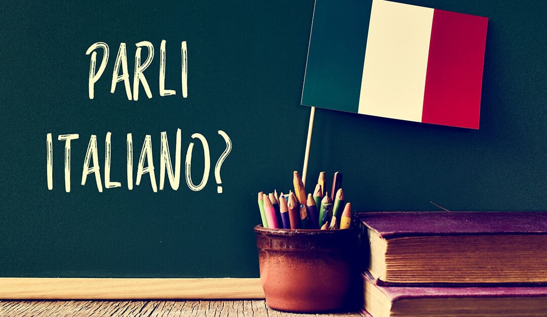 a chalkboard with the question parli italiano? do you speak Italian? written in Italian, a pot with pencils, some books and the flag of Italy, on a wooden desk