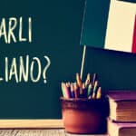 a chalkboard with the question parli italiano? do you speak Italian? written in Italian, a pot with pencils, some books and the flag of Italy, on a wooden desk