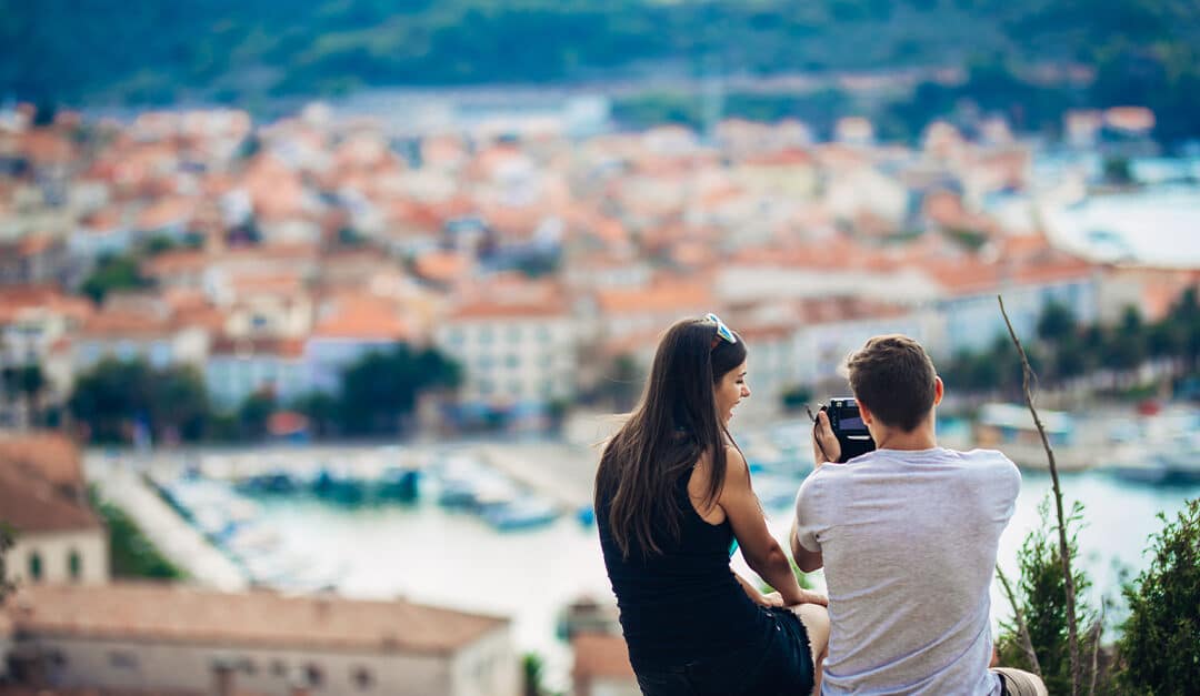 Young couple traveling and visiting Portugal sitting on a hill with city in the background, taking a picture with a camera. Blog Post - Basic Portuguese Phrases for Traveling to Portugal