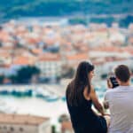 Young couple traveling and visiting Portugal sitting on a hill with city in the background, taking a picture with a camera. Blog Post - Basic Portuguese Phrases for Traveling to Portugal