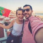 Handsome tourist couple take selfie photo with view of Lisbon, Portugal from Miradouro on sunny clear day holding Portugal flag - Cultural Tips when Traveling to Portugal