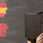 A young woman reading a Spanish text book sitting in front of a chalk board with Spanish and Mexican flags drawn in chalk on the board.