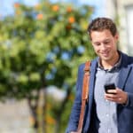 Young urban professional man using smart phone with Spanish chat abbreviations on street with trees in background