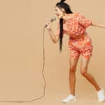 Young woman in orange clothes singing Portuguese songs into a microphone against a solid neutral background