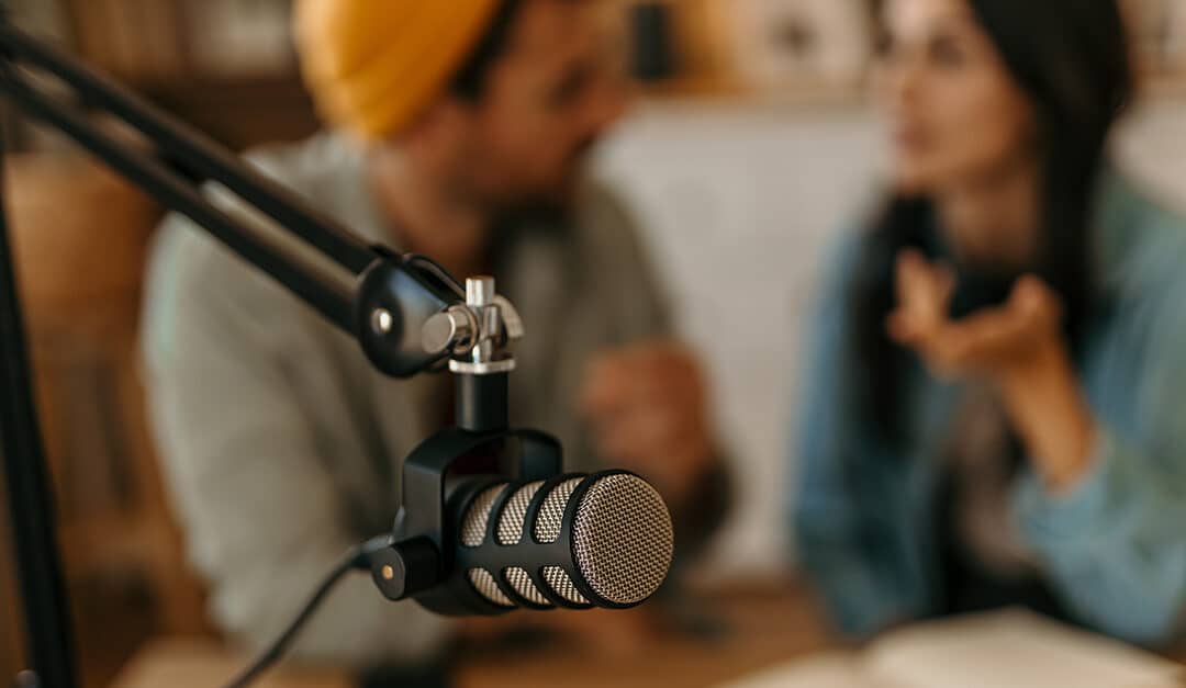 Two cheerful Portuguese people recording a podcast in a studio. Focus on the microphone in the foreground.