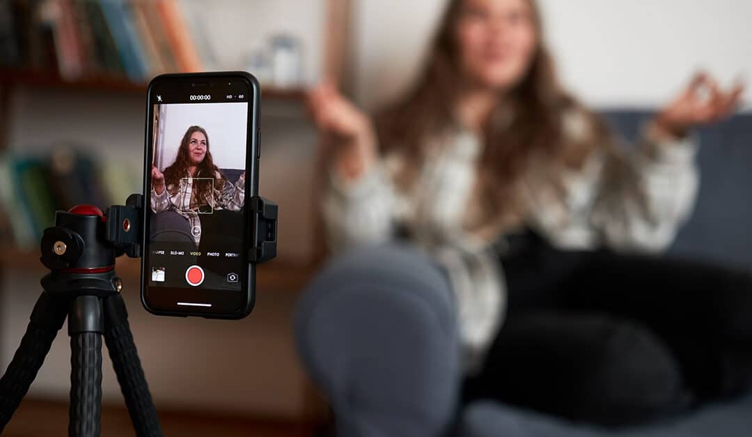 Selective focus on phone on tripod recording vlogger sitting on couch gesticulating and communicating with her followers | Learn Spanish with Vloggers