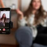 Selective focus on phone on tripod recording vlogger sitting on couch gesticulating and communicating with her followers | Learn Spanish with Vloggers