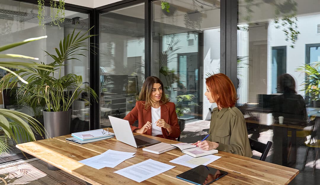 Two business women speaking Spanish in a glass walled conference room with many plants and ample sunlight.