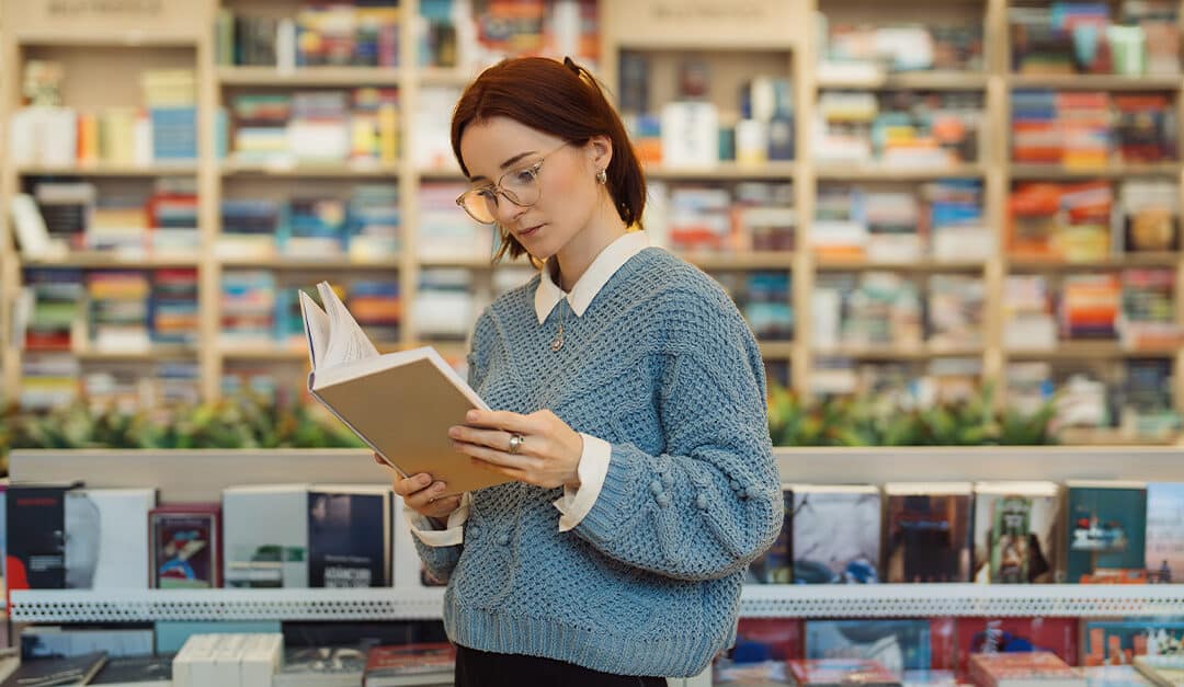 A focused young woman in glasses and a blue sweater reads a book while standing in a vibrant, well-stocked bookstore. The shelves are filled with a variety of books. Illustrating the blog topic of 3 Strategies to Learn Vocabulary in Another Language