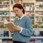 A focused young woman in glasses and a blue sweater reads a book while standing in a vibrant, well-stocked bookstore. The shelves are filled with a variety of books. Illustrating the blog topic of 3 Strategies to Learn Vocabulary in Another Language