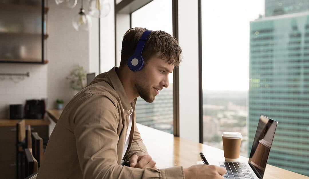 Young professional man with headphones and laptop, learning a new language while sat at a table or desk by a window of a high rise building