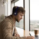 Young professional man with headphones and laptop, learning a new language while sat at a table or desk by a window of a high rise building