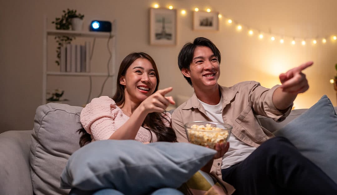 Young Asian couple on couch with popcorn watching and pointing to TV show, illustrating the blog topic Learn English for Daily Life by Watching “Friends”