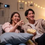 Young Asian couple on couch with popcorn watching and pointing to TV show, illustrating the blog topic Learn English for Daily Life by Watching “Friends”