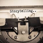 Close up black and white shot of a typewriter with "Storytelling..." typed on the sheet of paper, illustrating the blog topic "How Storytelling Helps You Learn a New Language Faster"