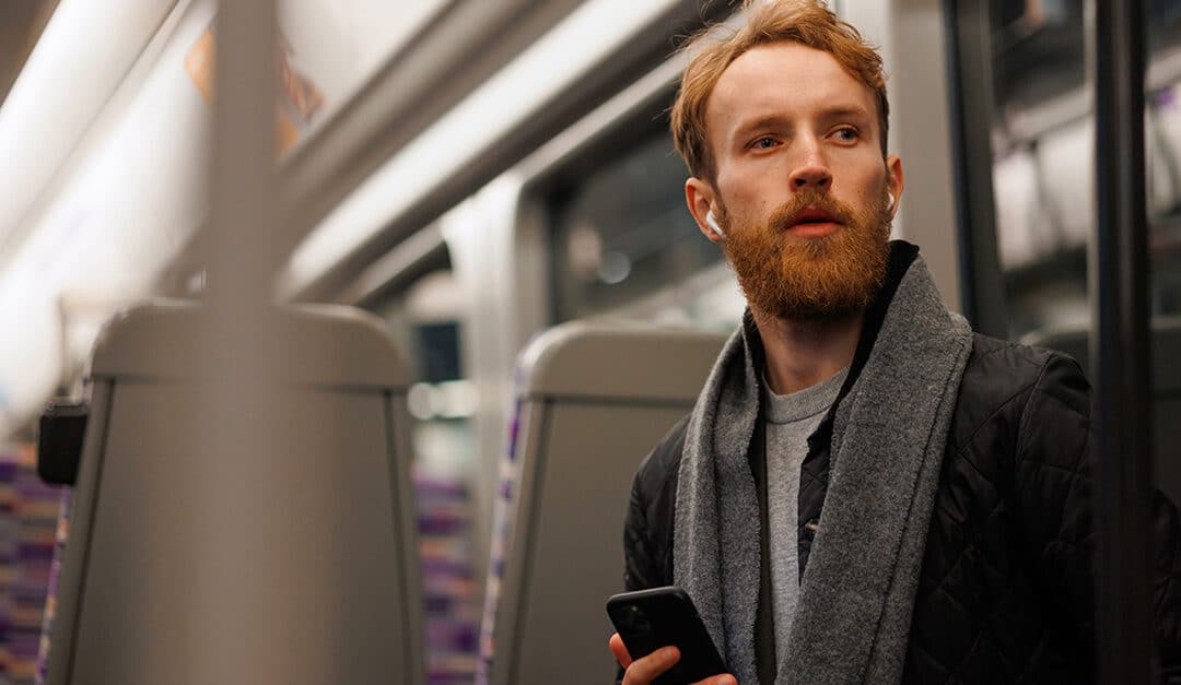 Red-headed, bearded professional male with earbuds, listening to a podcast on his public transportation commute, illustrating the blog topic "How to Learn a Second Language in Just 20 Minutes a Day"