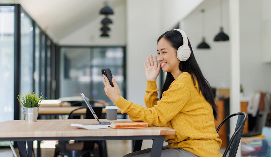 Young woman in yellow sweater, sitting at a table with laptop, headphones, and mobile, gesturing hello with her right hand while holding her phone in her left, illustrating the blog topic "How to Practice Speaking a Second Language: Top Resources to Boost Your Fluency"