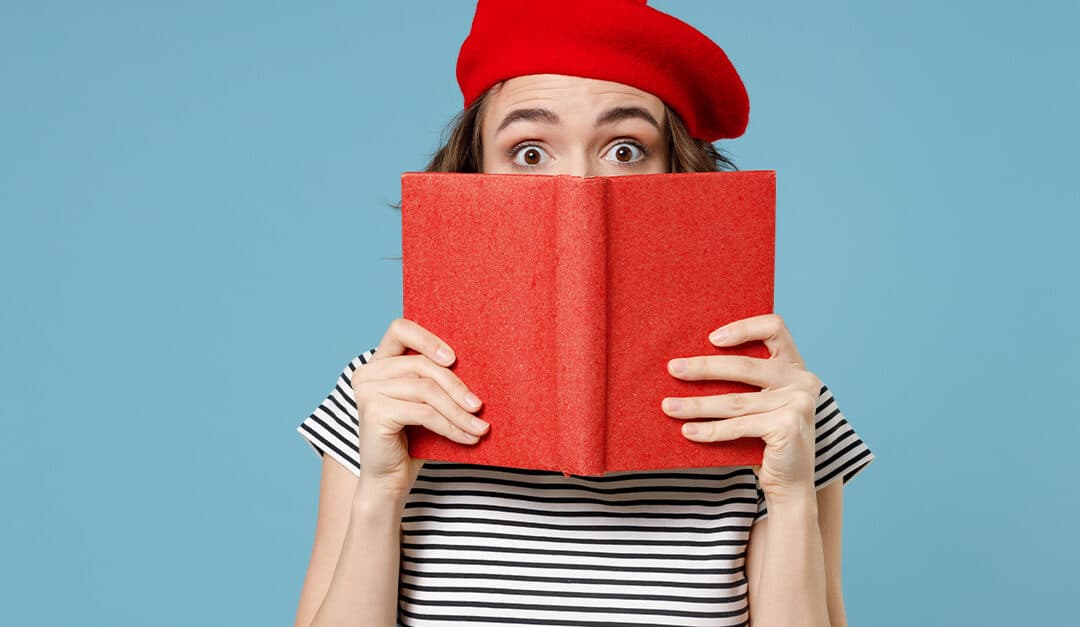 Young woman wearing French red beret and striped shirt holding a red book up to cover her face from the nose down on a blue background.