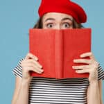 Young woman wearing French red beret and striped shirt holding a red book up to cover her face from the nose down on a blue background.