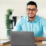 Young Arab male professional studying French online with a laptop and small French flag next to him on the desk