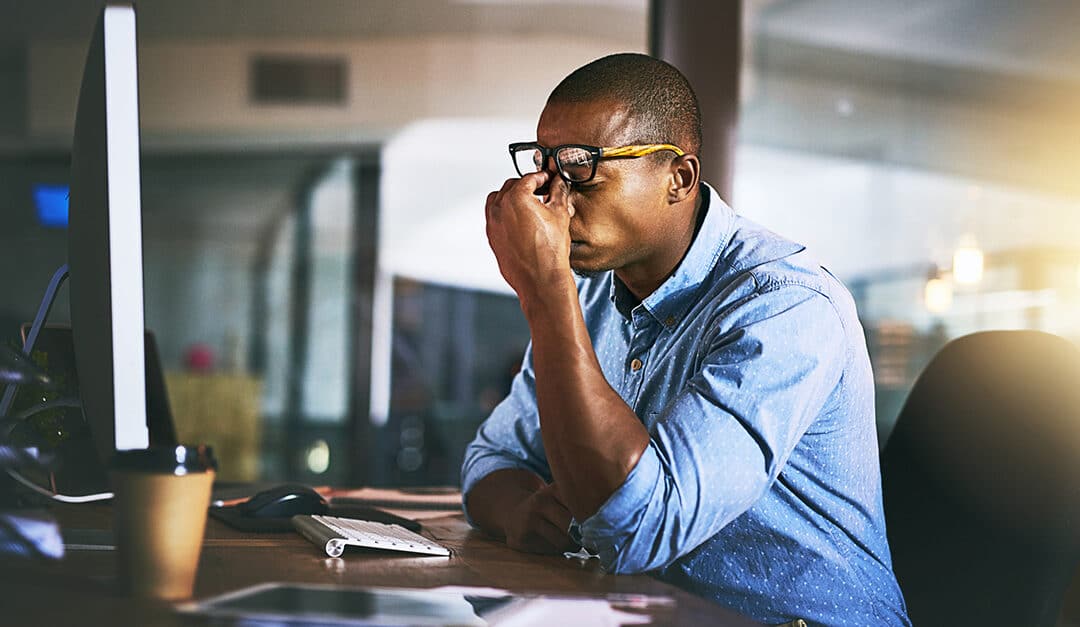 Stressed young professional male at a desk in an office, leaning on his desk and pinching the top of his nose with his eyes closed and glasses pushed up