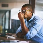 Stressed young professional male at a desk in an office, leaning on his desk and pinching the top of his nose with his eyes closed and glasses pushed up