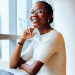 Smiling professional woman in glasses sitting at a desk by a window with a laptop
