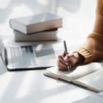 Woman at a desk writing in a notebook with books and a laptop nearby