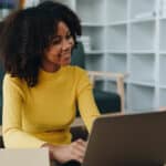 Woman in yellow sweater at a desk with a laptop, interacting with an online class