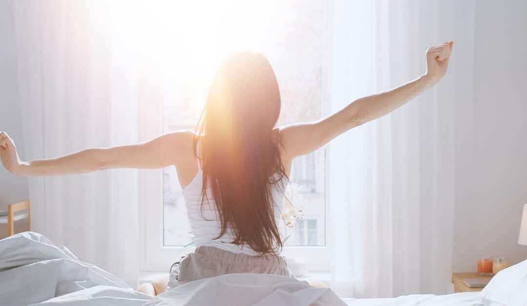 Young woman waking up in the morning and stretching while sitting on the bed and facing the sunrise in the window