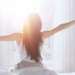 Young woman waking up in the morning and stretching while sitting on the bed and facing the sunrise in the window