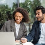 Young adult woman and man sitting outside looking at a laptop together and laughing